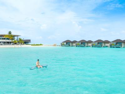 Klares türkisfarbenes Wasser mit einem Schwimmer und Strandbungalows vor weißem Sandstrand.