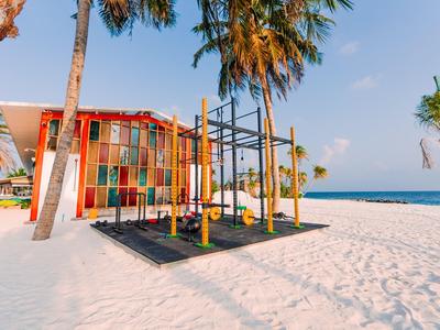 A colorful playground with climbing frames stands on white sand at the beach beside palm trees.