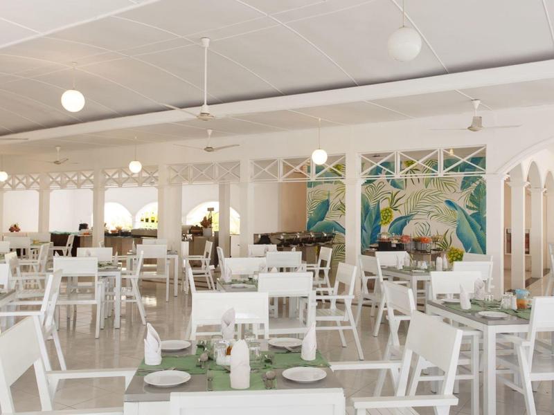 Světlá moderní restaurace s bílým nábytkem a tropickým nástěnným dekorem.