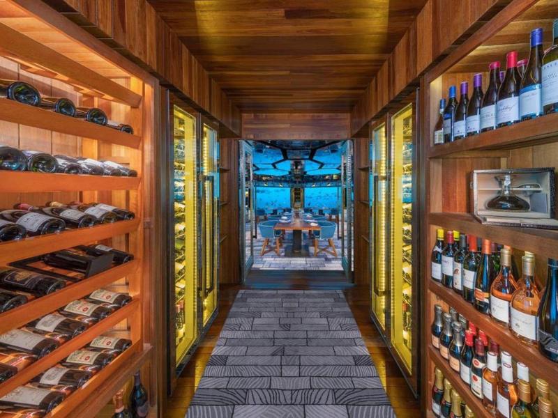 Pasillo con paredes de madera iluminadas y estantes de vino, vista a terraza exterior con mesa y sillas.