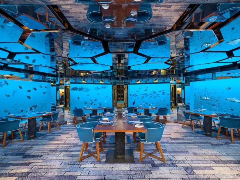Restaurante submarino con paredes transparentes y mesas con sillas frente a un arrecife de coral.