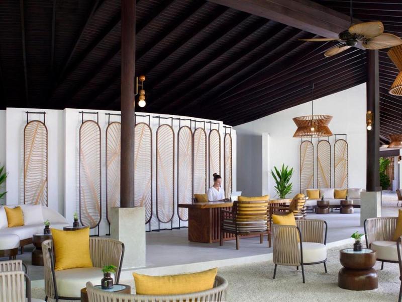 Elegancka hotelowa poczekalnia z białymi meblami, żółtymi poduszkami i drewnianym sufitem.