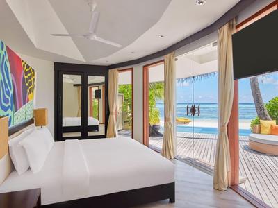 Lichte hotelkamer met groot bed, kleurrijke kunst en balkon met uitzicht op zwembad en oceaan.