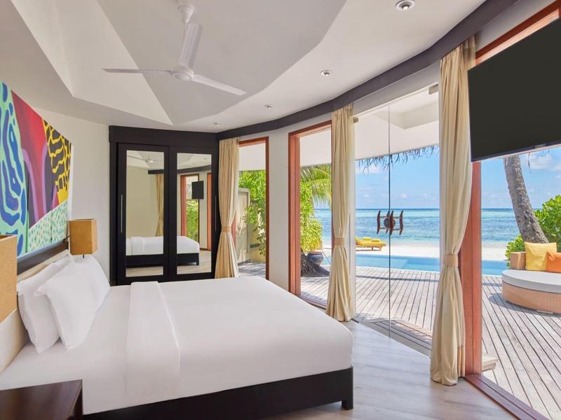 Lichte hotelkamer met groot bed, kleurrijke kunst en balkon met uitzicht op zwembad en oceaan.