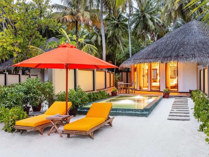 Tropische villa met zwembad, strandstoelen, oranje parasol, omgeven door palmbomen en groen.