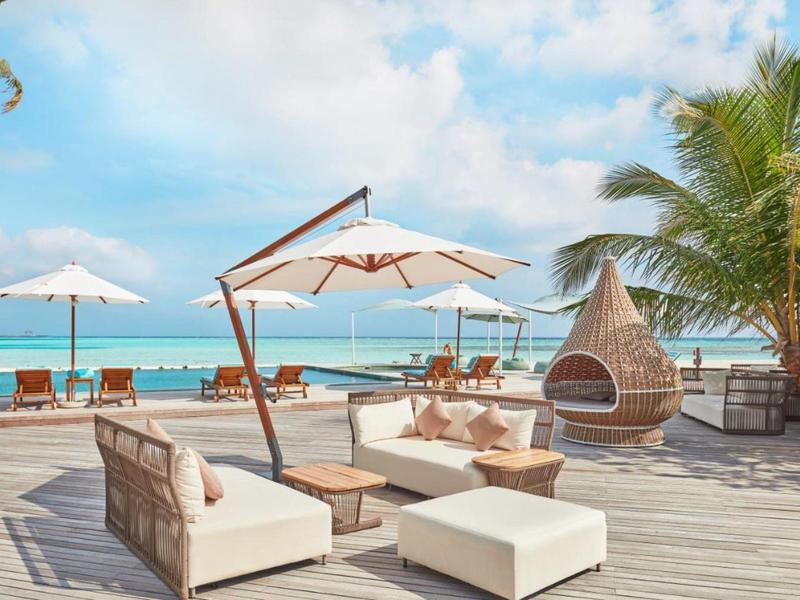 Loungegedeelte aan het strand met banken, parasols, palmbomen en uitzicht op de oceaan onder een blauwe lucht.