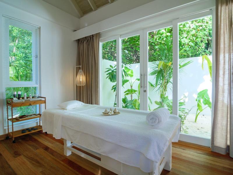 Helle Massagepraxis mit Holzfußboden, großer Fensterfront und weißem Massagetisch.