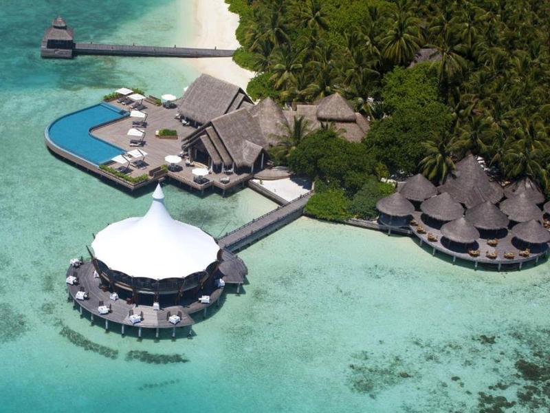 Aerial view of overwater bungalows and a pool at a tropical resort surrounded by clear turquoise water.