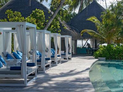 Area piscina con lettini bianchi e cuscini blu sotto palme in un resort tropicale.