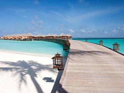 Dřevěná cesta vede nad tyrkysovou vodou k bungalovům nad vodou pod modrou oblohou.