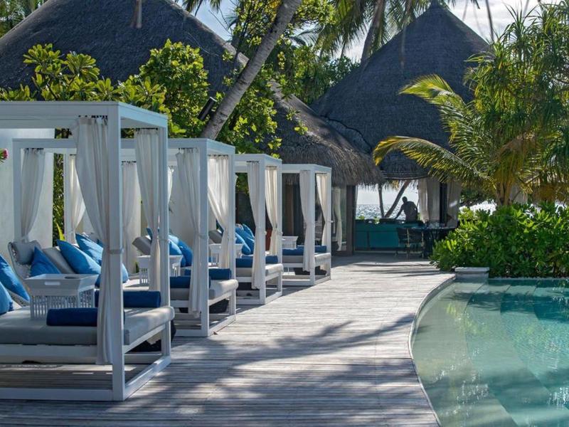 Area piscina con lettini bianchi e cuscini blu sotto palme in un resort tropicale.