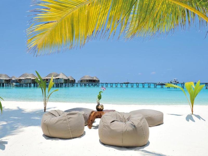 Spiaggia di sabbia bianca con pouf e palme sotto un cielo azzurro in un resort con bungalow sull'acqua