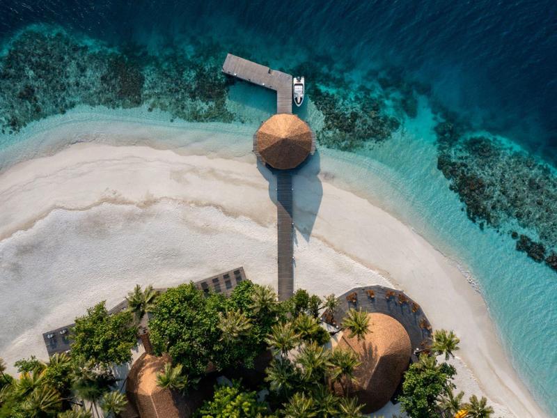 Aerial view of a tropical island with sandy beach, clear blue water, and wooden huts connected by a pier.