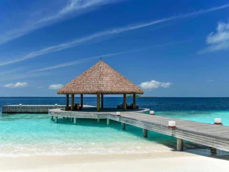 Wooden pier leads to thatched roof hut over clear blue tropical ocean water.