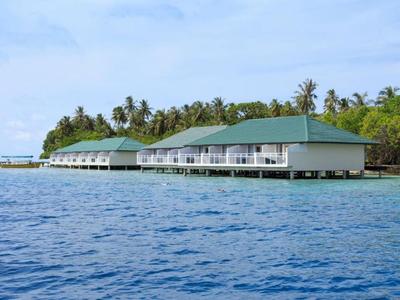 Wasserbungalows mit grünen Dächern auf Pfählen, umgeben von blauem Meer und Palmen im Hintergrund.