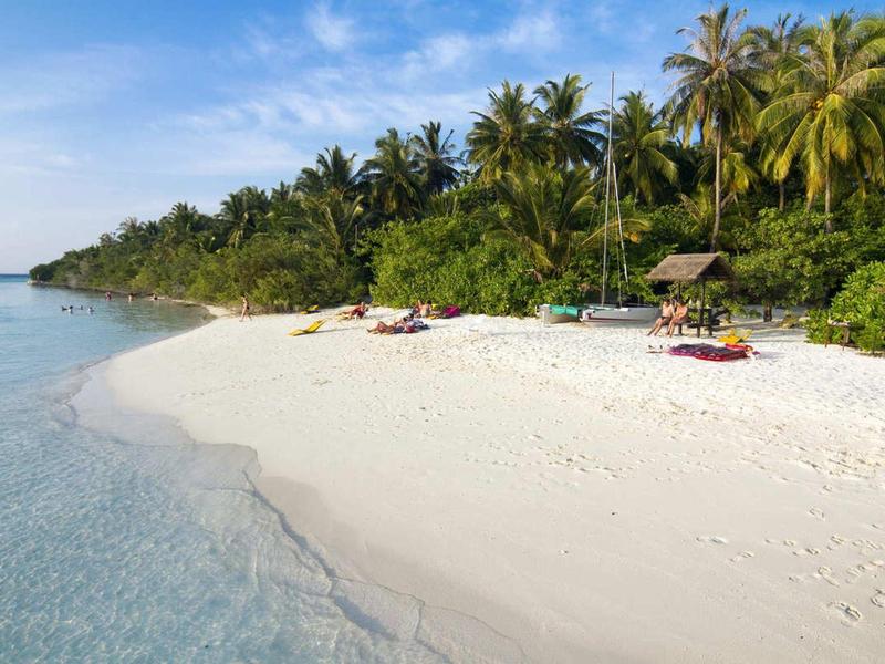 Bílá písečná pláž s průzračnou vodou a palmami v pozadí pod modrou oblohou.