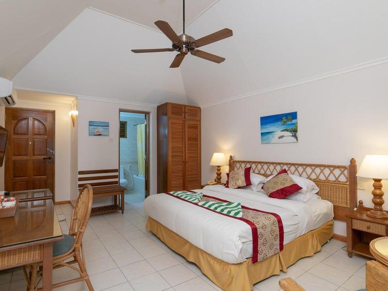 Prostorný hotelový pokoj s manželskou postelí, stropním ventilátorem a dřevěným psacím stolem.