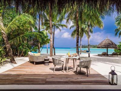 Seaside deck with chairs, table, lounge seat, palm trees, and ocean view under blue sky.