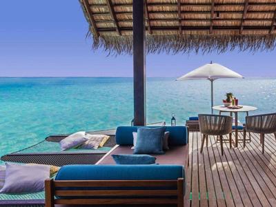 A wooden deck by clear blue ocean with cushioned seats, pillows, table, and umbrella under a thatched roof.