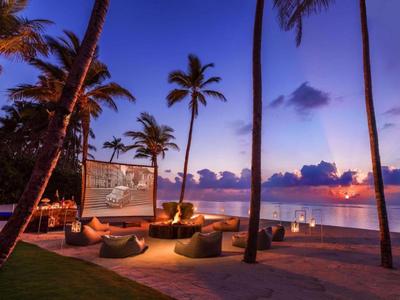 Lounge sulla spiaggia con pouf e schermo tra palme al tramonto sul mare.