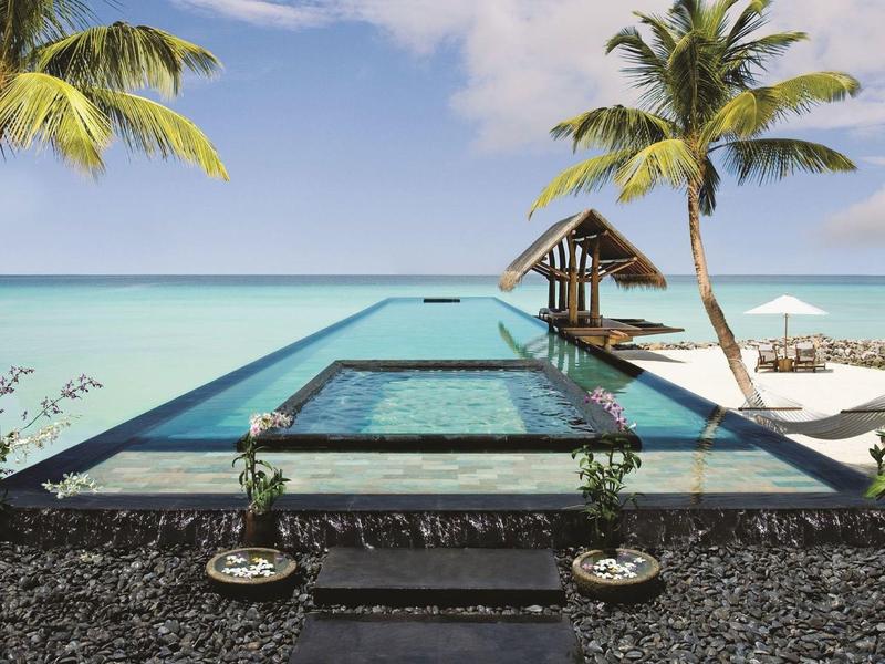 Infinity pool overlooking clear turquoise sea with palm trees and a small gazebo on a sandy beach.