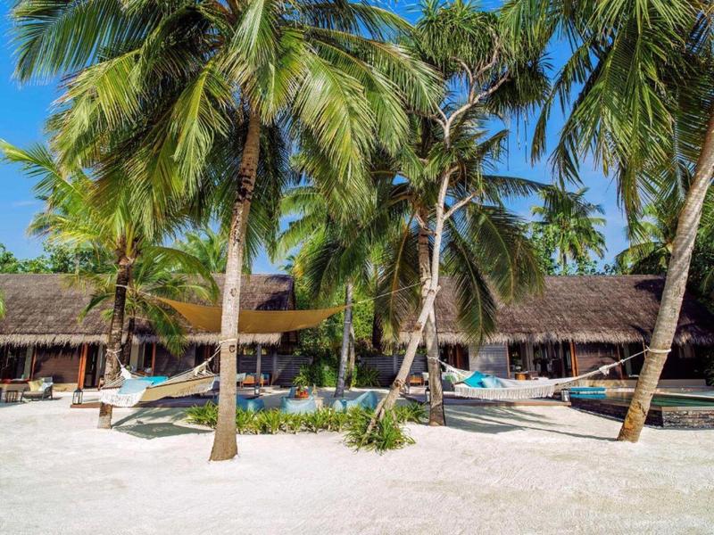 Tropical beach with palm trees, hammocks, and thatched roof huts under a clear sky.