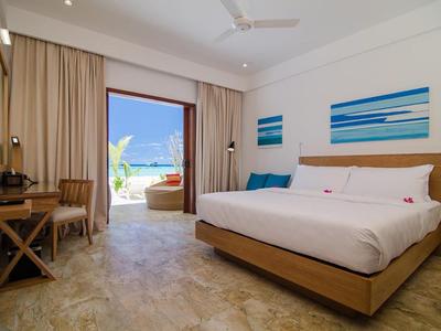 Helles Hotelzimmer mit großem Bett, Holzmöbeln und Blick auf das Meer durch eine offene Balkontür.