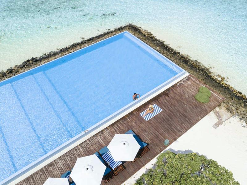 Ein rechteckiger Pool neben einem Strand mit zwei weißen Sonnenschirmen und einem Sonnendeck.