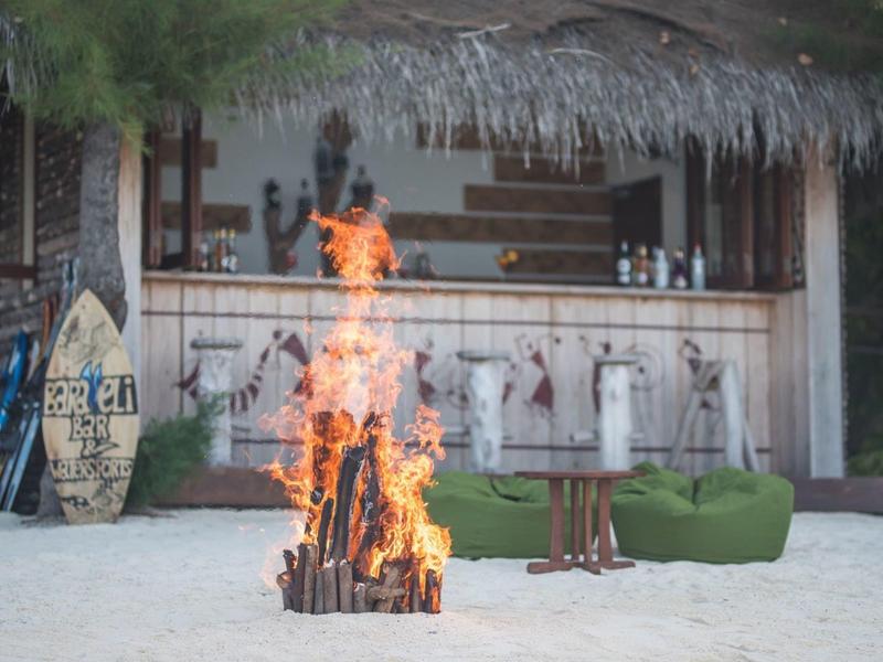 Lagerfeuer am Strand vor einer Bar mit Strohdach und grünen Sitzkissen im Sand.