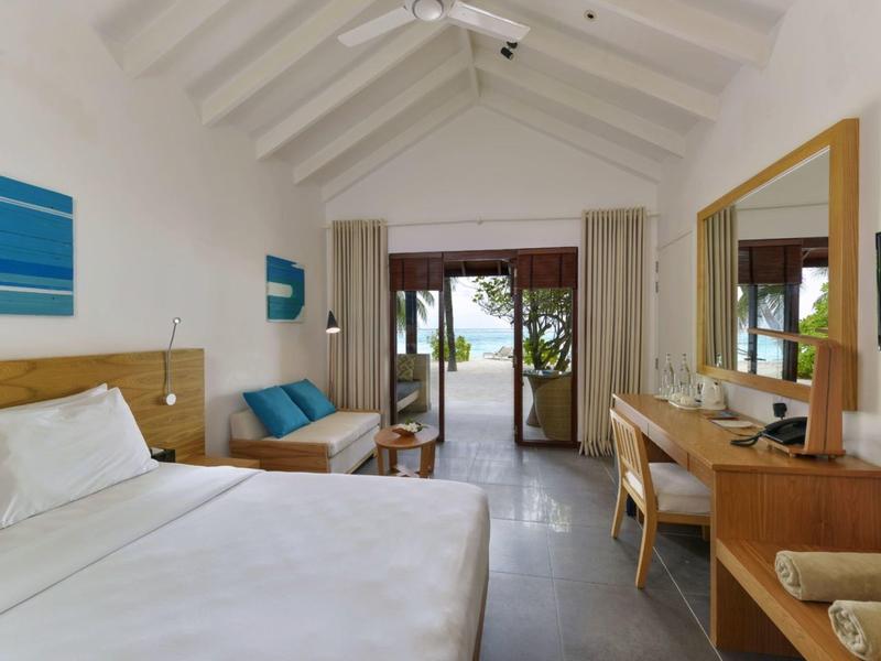 Helles Schlafzimmer mit weißem Bett, Holzmöbeln, blauem Sofa und Blick auf Terrasse mit Pool.