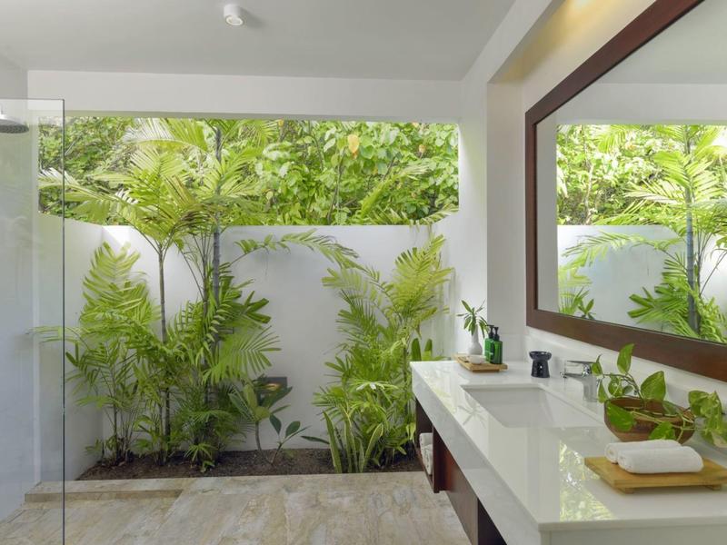 Helles Badezimmer mit großer Spiegelwand, weißem Waschtisch und grünen Pflanzen vor weißer Wand.