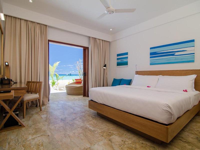 Helles Hotelzimmer mit großem Bett, Holzmöbeln und Blick auf das Meer durch eine offene Balkontür.