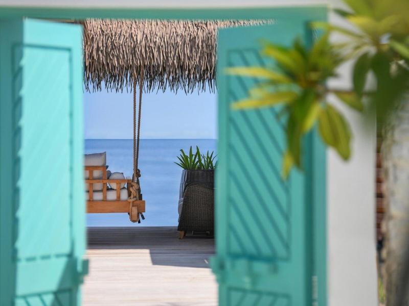 Widok przez otwarte turkusowe okiennice na drewnianą huśtawkę z widokiem na morze.