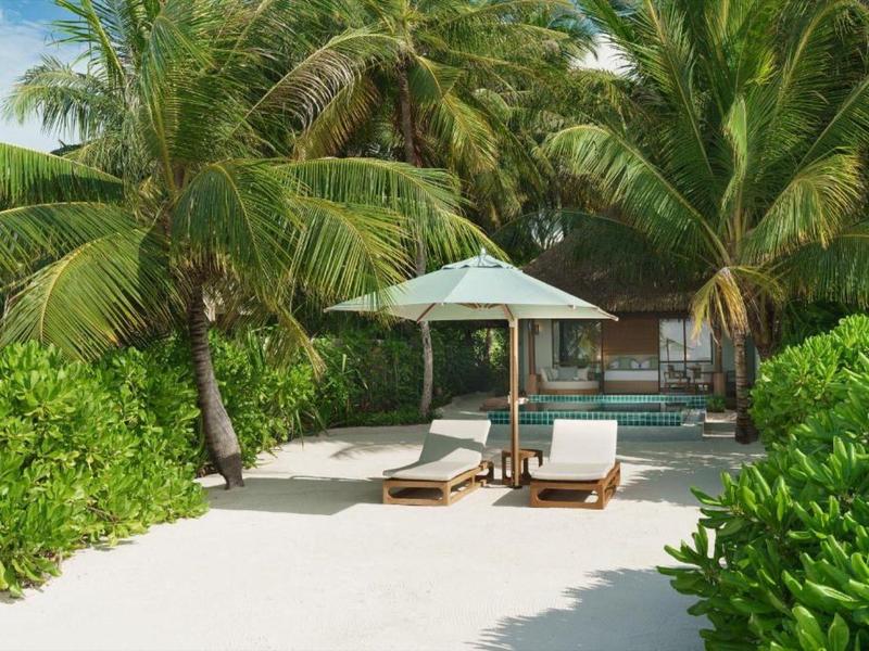 Deux chaises longues avec un parasol sur du sable blanc, entourées de palmiers et de plantes vertes.