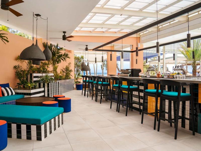 Helle Bar mit langen Theken, farbigen Sitzgelegenheiten und grünen Pflanzen im modernen Design.