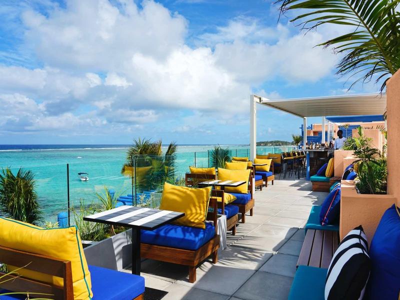 Dachterrasse mit bunten Sitzkissen, weißen Tischen, Palmen und Blick auf ruhiges türkisblaues Meer.