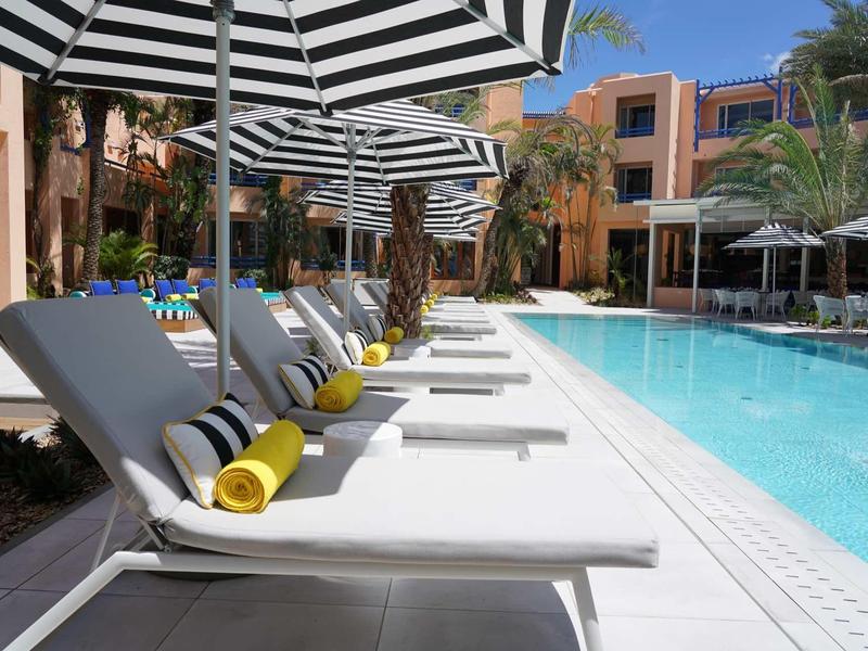 Poolbereich mit weißen Liegen, gestreiften Sonnenschirmen und gelben Handtüchern bei klarem Himmel.