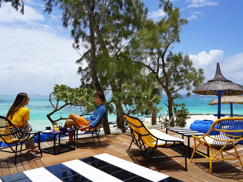 Strandterrasse mit Rattanmöbeln, zwei Personen, blaues Meer und Palmen im Hintergrund.