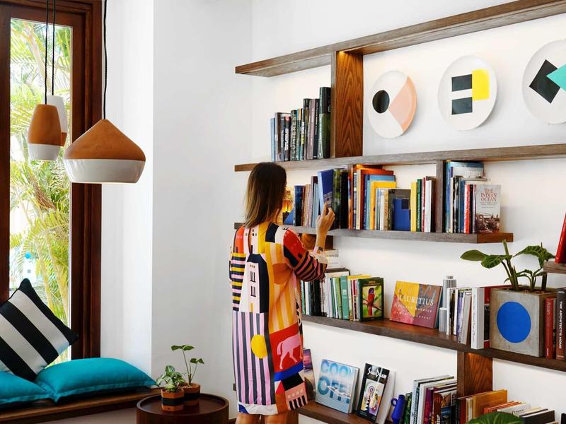 Frau in buntem Kleid steht vor Holzregal mit Büchern, neben Fenster und blauem Sitzkissen.