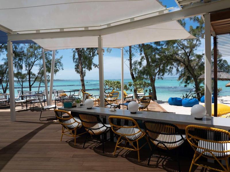 Offener Strandbereich mit Tischen und Stühlen, Palmen und Meer im Hintergrund, sonnig und einladend.