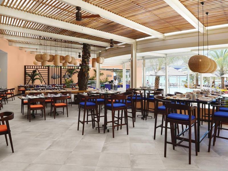 Modernes Restaurant mit Holzdecke, farbigen Stühlen und großer Fensterfront zu einer Außenterrasse.