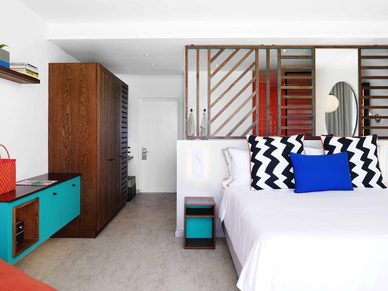 Modern eingerichtetes Hotelzimmer mit Holzmöbeln, weißem Bett und bunten Akzenten.