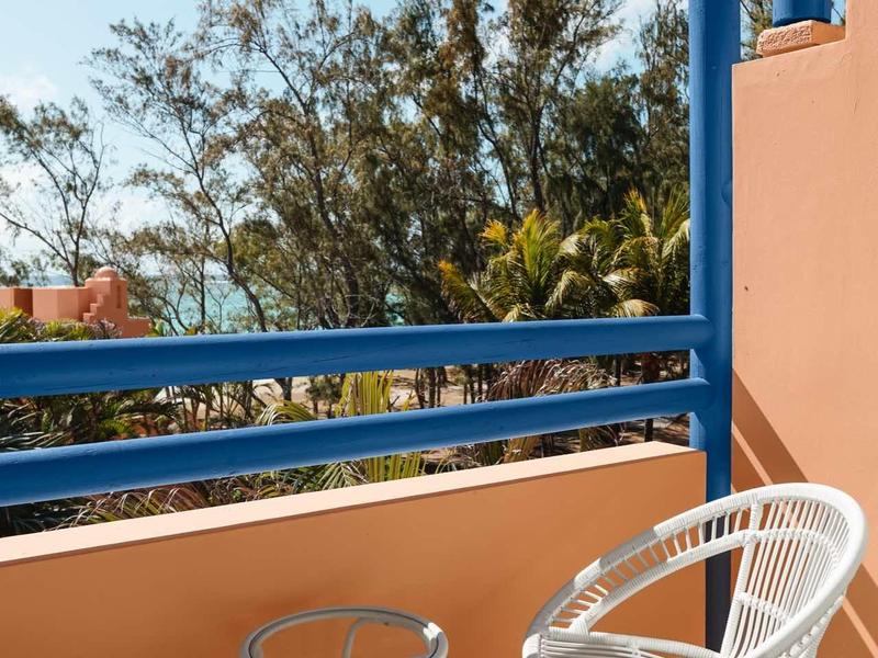 Balkon mit blauen Geländern, zwei weißen Stühlen und kleinem Tisch mit Blick auf Bäume und Meer.