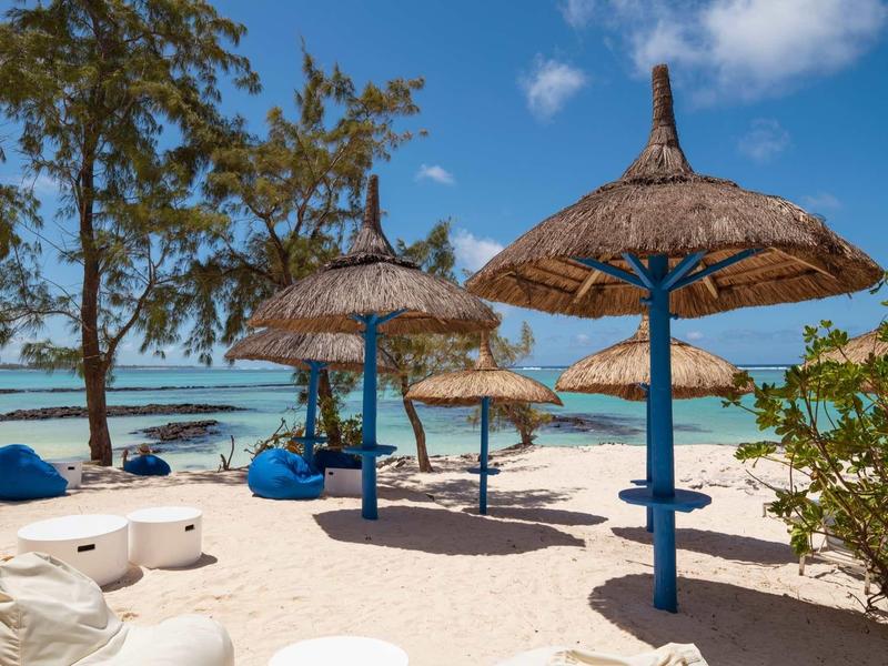 Strand mit weißen Sand, blauen Sitzsäcken und strohgedeckten Sonnenschirmen unter blauem Himmel.