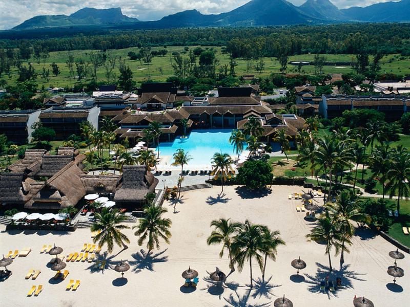 Ein Strandresort mit Palmen, Sonnenschirmen, Pool und Bergen im Hintergrund unter bewölktem Himmel.