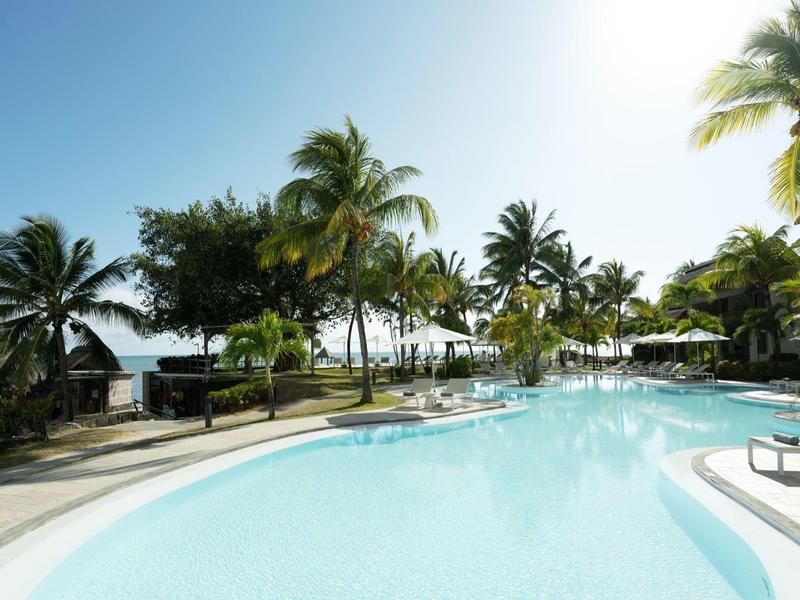 Großer Pool mit klarem Wasser, umgeben von Palmen, Sonnenschirmen und blauen Himmel.