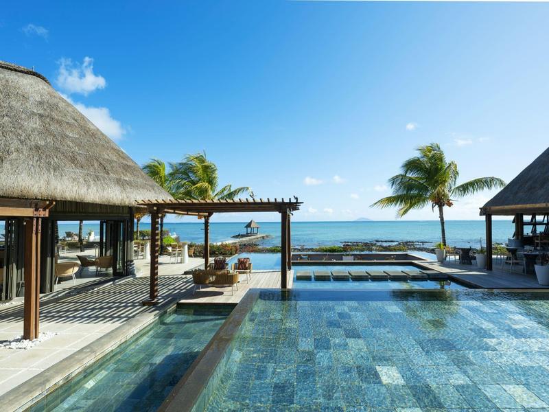 Luxuriöse Poolanlage mit klarem Wasser, Palmen und offenem Blick aufs Meer unter blauem Himmel.