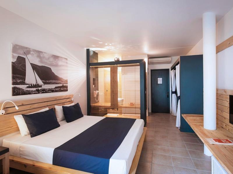 Chambre d'hôtel moderne avec lit double, douche en verre, détails en bois et décoration murale maritime.