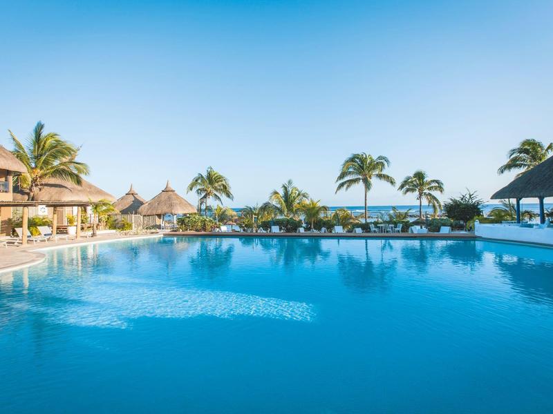 Grande piscine d'hôtel avec transats et palmiers sous un ciel bleu clair