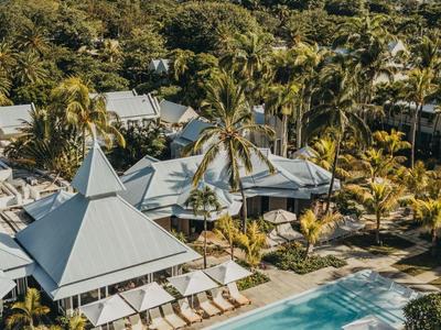 Blick auf einen Pool mit Liegen, umgeben von Palmen und bungalowartigen Unterkünften.